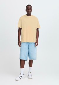Beige oversized t-shirt met korte mouwen, gecombineerd met lichtblauwe denim shorts en witte sneakers met zwarte strepen. Minimalistisch ontwerp.