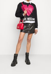Schwarzer Sweatshirt mit pinkem Herz-Graphic und "LOVE MOSCHINO" Text, kombiniert mit einem schwarzen Leder-Minirock mit Knöpfen an der Vorderseite. Eine rote gesteppte Handtasche und schwarze Schnürstiefel runden das Outfit ab.