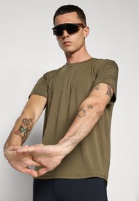 T-shirt verde oliva em tecido macio com mangas curtas. Modelo usa óculos de sol pretos, exibindo tatuagens nos braços e um fundo neutro.