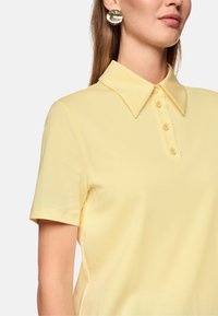 Blassgelbes Polohemd mit klassischem Kragen und kurzen Ärmeln, ausgestattet mit einer Knopfleiste mit mehreren Knöpfen. Glatte Textur.