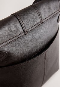 Geantă maro din piele, cu suprafață texturată, accente cusute și un buzunar frontal. Formă rectangulară compactă cu închidere sigură.