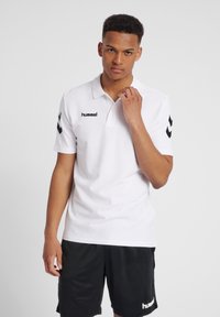 Witte polo shirt gemaakt van katoen met een kraag, voorzien van een zwart hummel logo en zwarte chevronaccenten op de mouwen. Gekozen in combinatie met zwarte shorts.