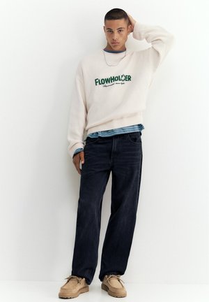 Ung mand iført en hvid Flowholder sweater, mørke jeans, beige sko, stående op ad en hvid væg med en hånd på hovedet.