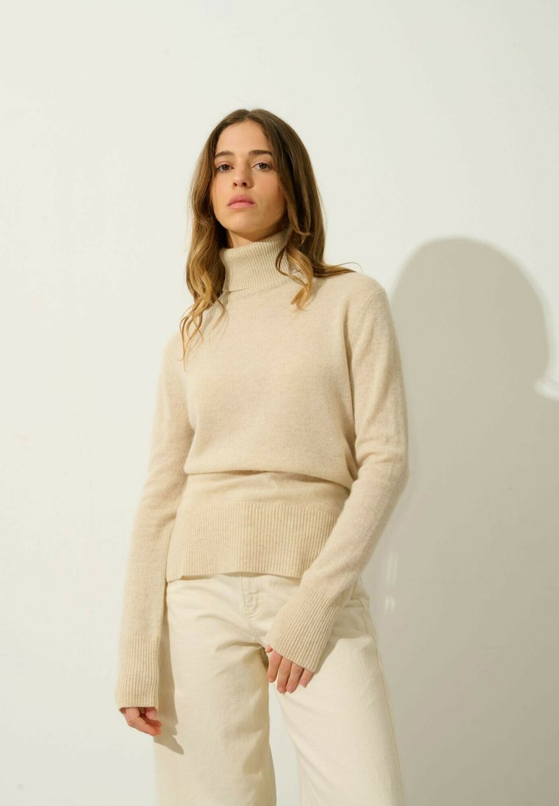 Beżowy sweter z golfem wykonany z miękkiego materiału, z prążkowanymi detalami u dołu i na mankietach, zestawiony z jasnymi spodniami.