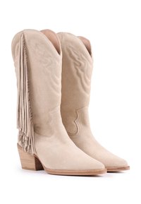 Botas de vaquero de ante beige con detalles de flecos en la parte trasera, puntera afilada y un tacón de madera apilada. Costuras decorativas a lo largo de los lados.