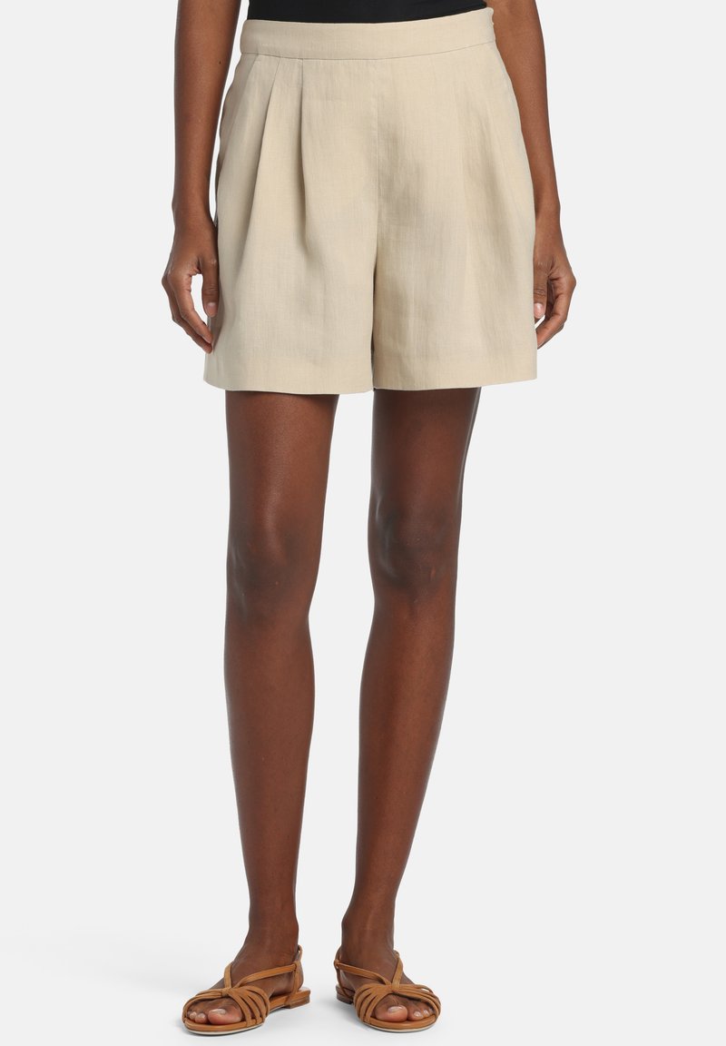 Beige plisserede shorts båret af en person med mørk hud, kombineret med brune flade sandaler med stropper mod en ensfarvet baggrund.