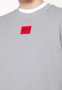 Sudadera gris con cuello redondo y un parche rectangular rojo que lleva la palabra "HUGO" en texto negro. Tejido suave con puños de canalé.