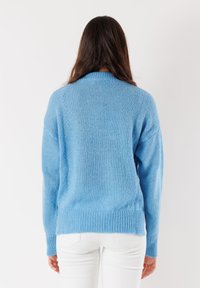 Donna con lunghi capelli castani che indossa un maglione blu in maglia e pantaloni bianchi, mostrata di spalle su uno sfondo bianco.