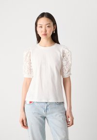 Tricou alb cu mâneci din dantelă umflată. Material de bumbac moale, decolteu rotund, croială lejeră și textură detaliată de dantelă florală pe mâneci.