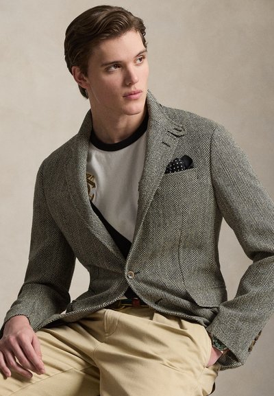 Νεαρός άνδρας καθιστός, φοράει σακάκι ψαροκόκαλο με μαντηλάκι τσέπης που έχει βούλες, γραφικό T-shirt, μπεζ παντελόνι και ρολόι χειρός, κοιτάζει πλάγια.