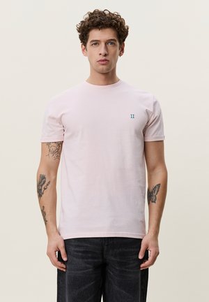 Jeune homme aux cheveux bouclés porte un t-shirt rose clair avec un petit logo bleu sur la poitrine et un jean noir, debout devant un fond clair uni.