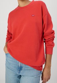 Sudadera roja de cuello redondo con un pequeño parche del logo de Levi's en el pecho. Cuello redondo, corte relajado y puños acanalados. Combinada con jeans de mezclilla azul claro.