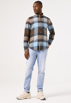 Chemise à carreaux dans des tons marron, bleu et crème, design boutonnière, associée à un jean bleu clair et des baskets beiges, présentée debout.