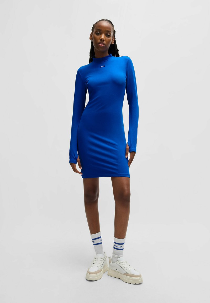 Blauwe lange mouwen jurk met een hoge kraag, gladde textuur en een getailleerd ontwerp. Gecombineerd met witte sneakers en kniekousen met blauwe accenten.
