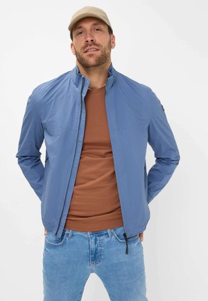 Blue lightweight jacket with stand-up collar, front zipper, and two side pockets, worn over a brown shirt and paired with blue jeans.