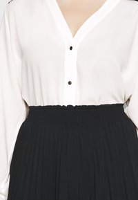 Primo piano di una persona che indossa una blusa bianca a collo a V con due bottoni, abbinata a una gonna nera plissettata in vita.