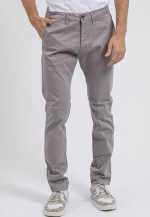 Homme portant un pantalon slim gris clair et des baskets blanches avec des accents gris, debout avec une main dans la poche devant un fond uni.