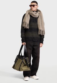 Pull en maille à motifs noirs et gris, écharpe à franges beige, pantalon noir et lunettes de soleil. Sac olive avec des anses noires, baskets blanches.