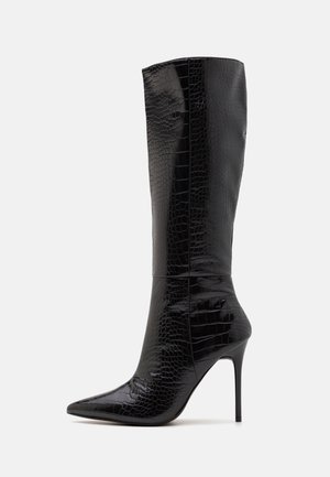 Bota de tacón stiletto negra hasta la rodilla con punta afilada y textura de cocodrilo, mostrada de lado sobre un fondo blanco.