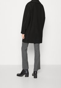 Cappotto oversize nero con cuciture a contrasto, rever inclinati; abbinato a pantaloni grigi a quadri e stivaletti neri, texture liscia.