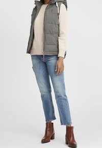 Grau gesteppte Weste mit Kapuze, beige Sweatshirt darunter, hellblaue Jeans und braune Ankle Boots. Ausgestattet mit Reißverschluss, Zugbändern und Taschenakzenten.