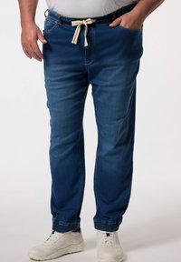 Pantalones jogger de mezclilla en azul oscuro, con cintura elástica y cordón, bolsillos laterales y puños ajustados. Llevados con zapatillas blancas.