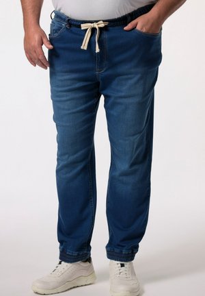 Pantalones jogger de mezclilla en azul oscuro, con cintura elástica y cordón, bolsillos laterales y puños ajustados. Llevados con zapatillas blancas.