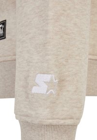 Manche de sweat-shirt beige avec un "S" brodé blanc et un logo étoile près du poignet côtelé, ainsi qu'une petite étiquette en tissu noire carrée en arrière-plan.