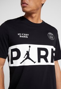 Hombre que lleva una camiseta negra con el texto "ICI C’EST PARIS", el logo del Paris Saint-Germain y un gran texto "PARIS" que incorpora la silueta de un jugador de baloncesto.