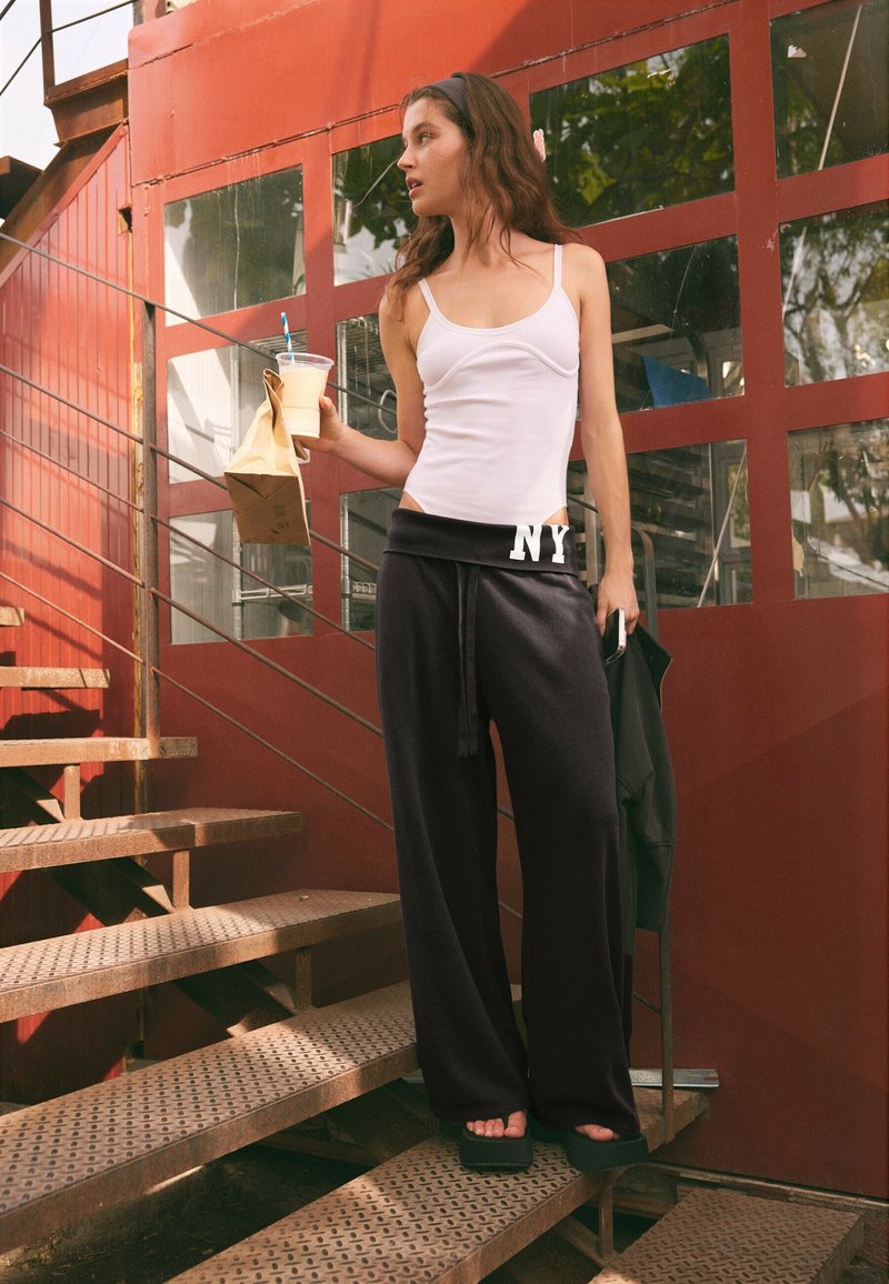 Jeune femme en débardeur blanc et pantalon noir tenant une boisson et un sac en papier, debout sur un escalier métallique à l'extérieur d'un bâtiment rouge.