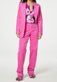 Completo in corduroy rosa con giacca su misura e pantaloni abbinati. Sotto, una blusa multicolore con motivi astratti. Scarpe rosa glitterate.