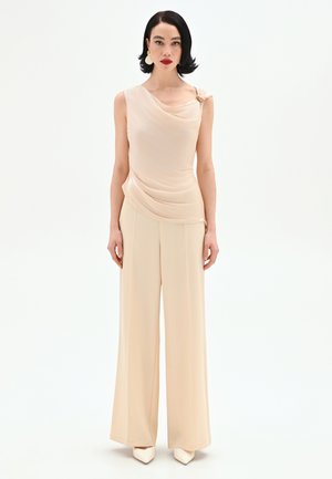 Donna con capelli neri e rossetto rosso che indossa un top beige senza maniche drappeggiato, pantaloni beige a gamba larga, scarpe bianche a punta e grandi orecchini.