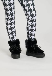 Black faux fur boots with a high cuff, featuring black laces, platform soles, and textured rubber outsoles, paired with black-and-white houndstooth leggings.