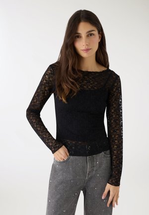 Donna con capelli lunghi castani che indossa una maglietta di pizzo nera a maniche lunghe e jeans grigi a vita alta con piccole macchie di vernice bianche, in piedi con una mano in tasca.