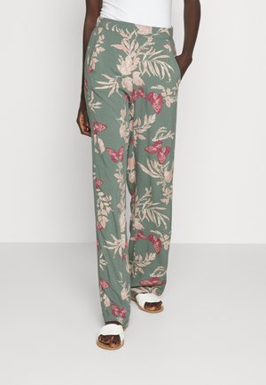 Woman wearing green wide-leg pants with pink floral and butterfly print, paired with white sandals and a white top.