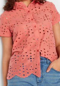 Korale różowy krótki rękaw koszula zapinana na guziki z haftem w kształcie oczek, noszona włożona w niebieskie dżinsy.