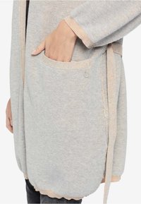 Grijze gebreide cardigan met een licht perzikkleurige accent, voorzien van een zijzak, striksluiting en textuurdetail in de stof.
