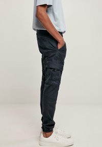Personne portant un pantalon cargo noir avec les chevilles retroussées, des baskets blanches et une chemise bleu clair, vue de profil, une main dans la poche.