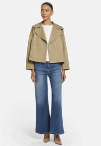 Vrouw in een beige cropped double-breasted jacket, witte top, blauwe flared jeans en beige hoge hakken, staande voor een witte achtergrond.