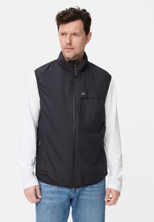 Mężczyzna w czarnej kamizelce bez rękawów zapinanej na zamek, noszący białą, długorękawową koszulę i niebieskie dżinsy, patrzący lekko na bok.