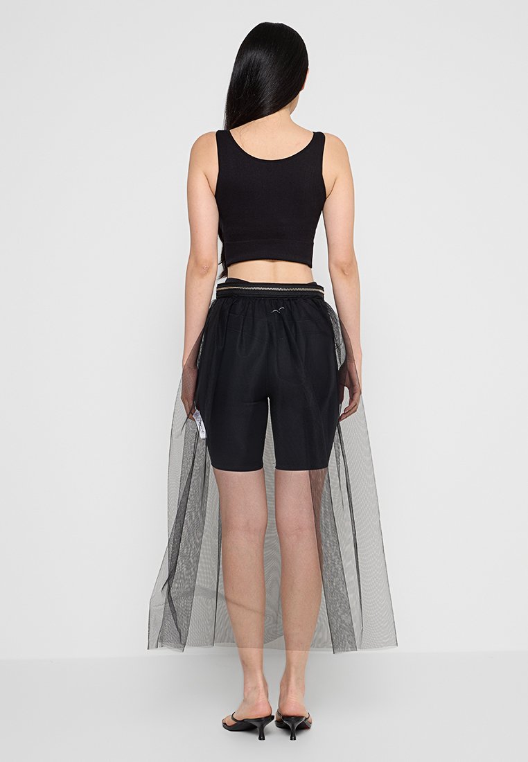 Donna con lunghi capelli neri indossa un crop top nero senza maniche, pantaloni shorts neri con sovrapposizione di gonna in rete trasparente e sandali neri con tacco, in piedi con le spalle rivolte verso la fotocamera.