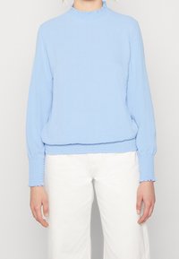 Top azzurro chiaro a maniche lunghe con collo alto arricciato e polsini smock, caratterizzato da una vestibilità rilassata e un tessuto testurizzato, abbinato a pantaloni bianchi.