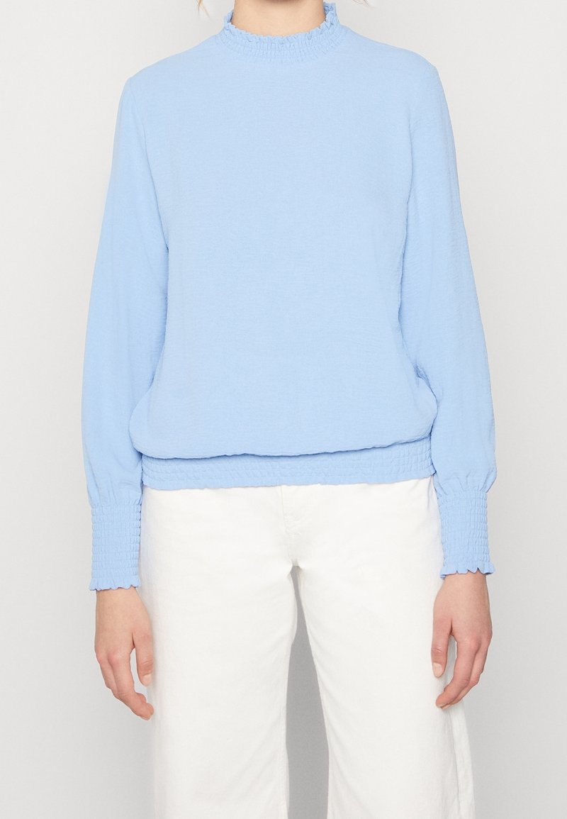 Top azzurro chiaro a maniche lunghe con collo alto arricciato e polsini smock, caratterizzato da una vestibilità rilassata e un tessuto testurizzato, abbinato a pantaloni bianchi.