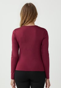Maglietta a maniche lunghe bordeaux con scollo rotondo e silhouette aderente. Tessuto morbido con una leggera lucentezza. Design minimalista senza decorazioni.