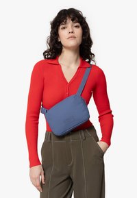 Slingväska i mjukt blått tyg med en texturerad etikett. Har en smal rektangulär form och justerbar rem. Bärs i en crossbody-stil.