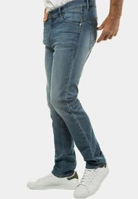 Vaqueros de denim azul con un corte ajustado, que cuentan con cinco bolsillos, un ligero desgaste y costuras en contraste. Combinados con zapatillas blancas.