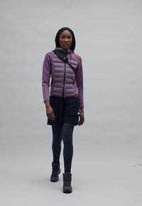 Jachetă izolată purpurie cu închidere cu fermoar, pantaloni scurți negri, colanți închiși la culoare și bocanci de drumeție gri. Modelul merge pe un fundal uniform.