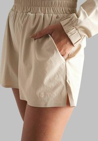 Short beige avec une ceinture élastique froncée et des poches latérales, avec un logo discret embossé sur le tissu. Matière lisse et légère.
