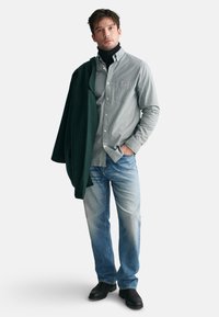 Grau-weiß kariertes Hemd, dunkler Rollkragenpullover, grüne Jacke über die Schulter drapiert, hellblaue Jeans und schwarze Schuhe. Lässiger Look.