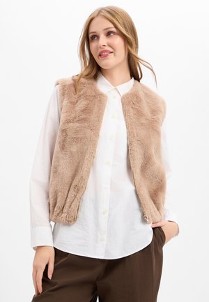 Giovane donna che indossa un gilet beige peloso sopra una camicia bianca con bottoni e pantaloni marroni, in piedi con una mano in tasca davanti a uno sfondo bianco.